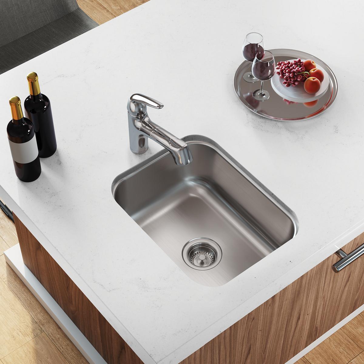 Stainless steel undermount bar sink installed in white countertop with chrome faucet and drain strainer visible.