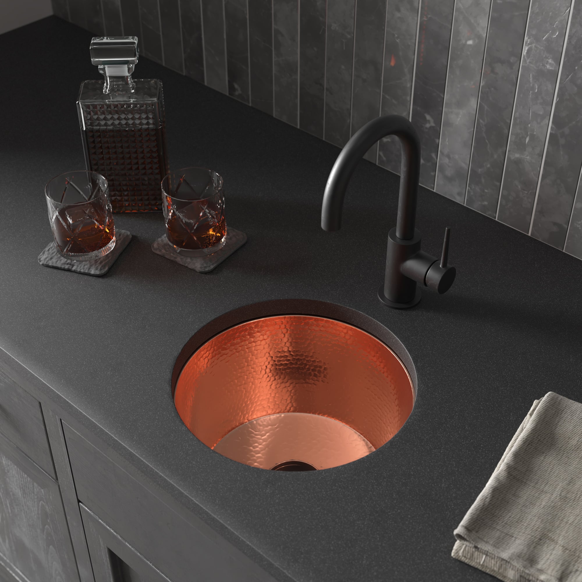Polished copper round undermount sink with black curved faucet on dark countertop next to whiskey glasses and decanter.