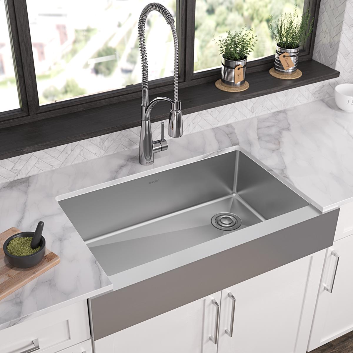 Stainless steel farmhouse sink with single bowl and apron front, installed in marble countertop with chrome faucet.
