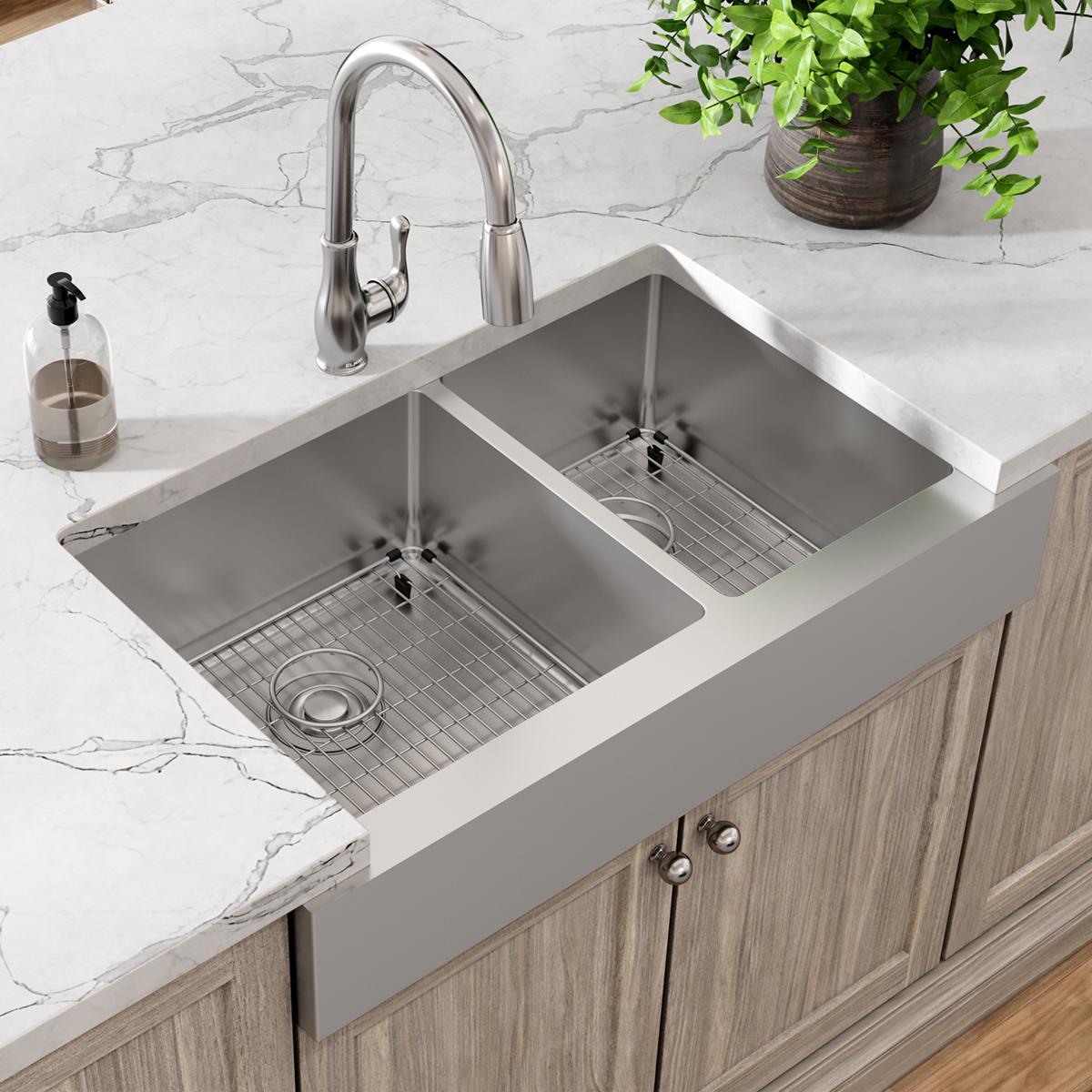 Stainless steel farmhouse sink with two unequal bowls, pull-down faucet, and grid strainers installed in white countertop.