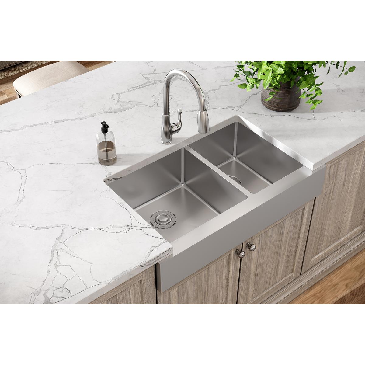 Stainless steel double bowl farmhouse sink with unequal sized compartments installed in white countertop with faucet.