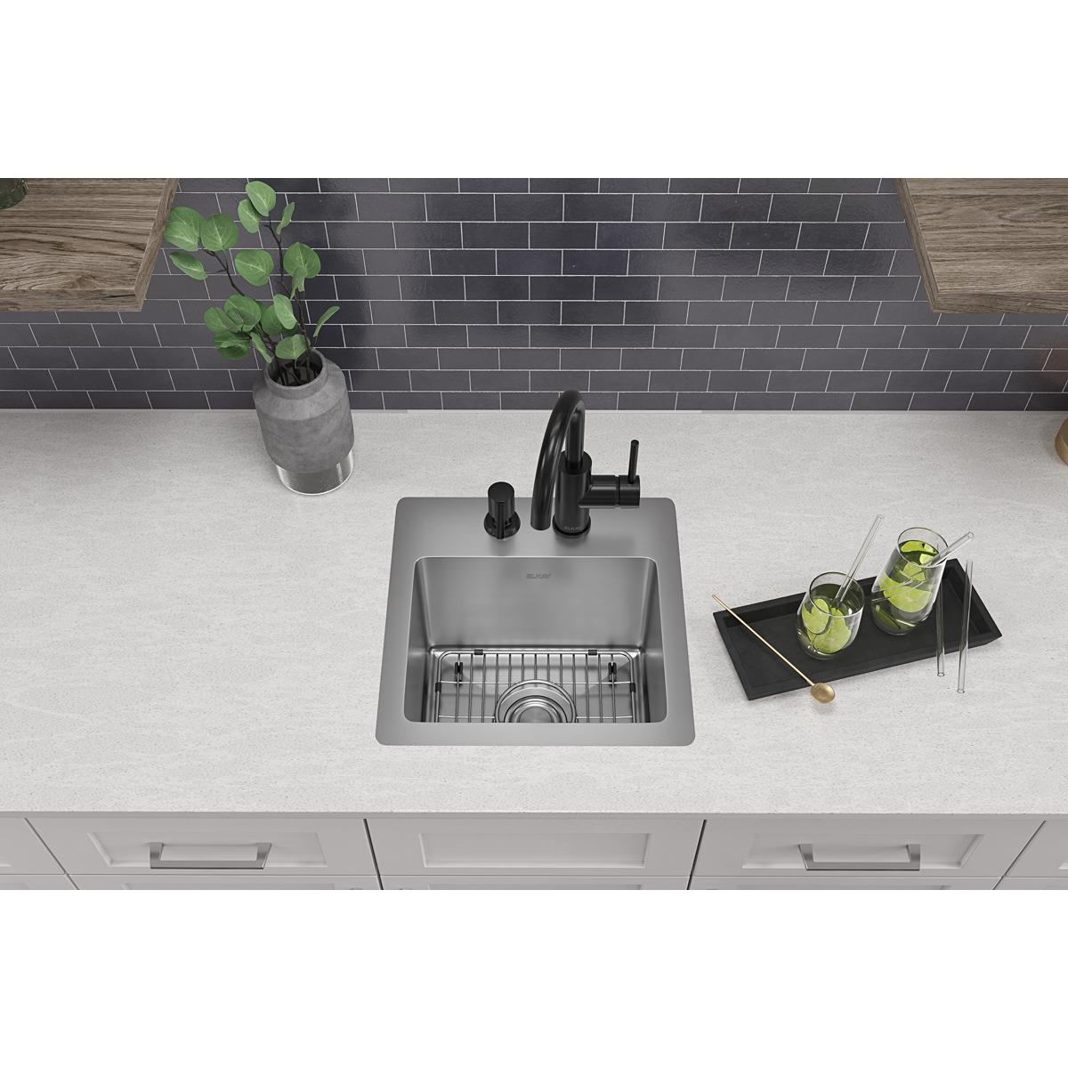 Stainless steel single bowl bar sink with two faucet holes, black faucet, and grid insert installed in white countertop.