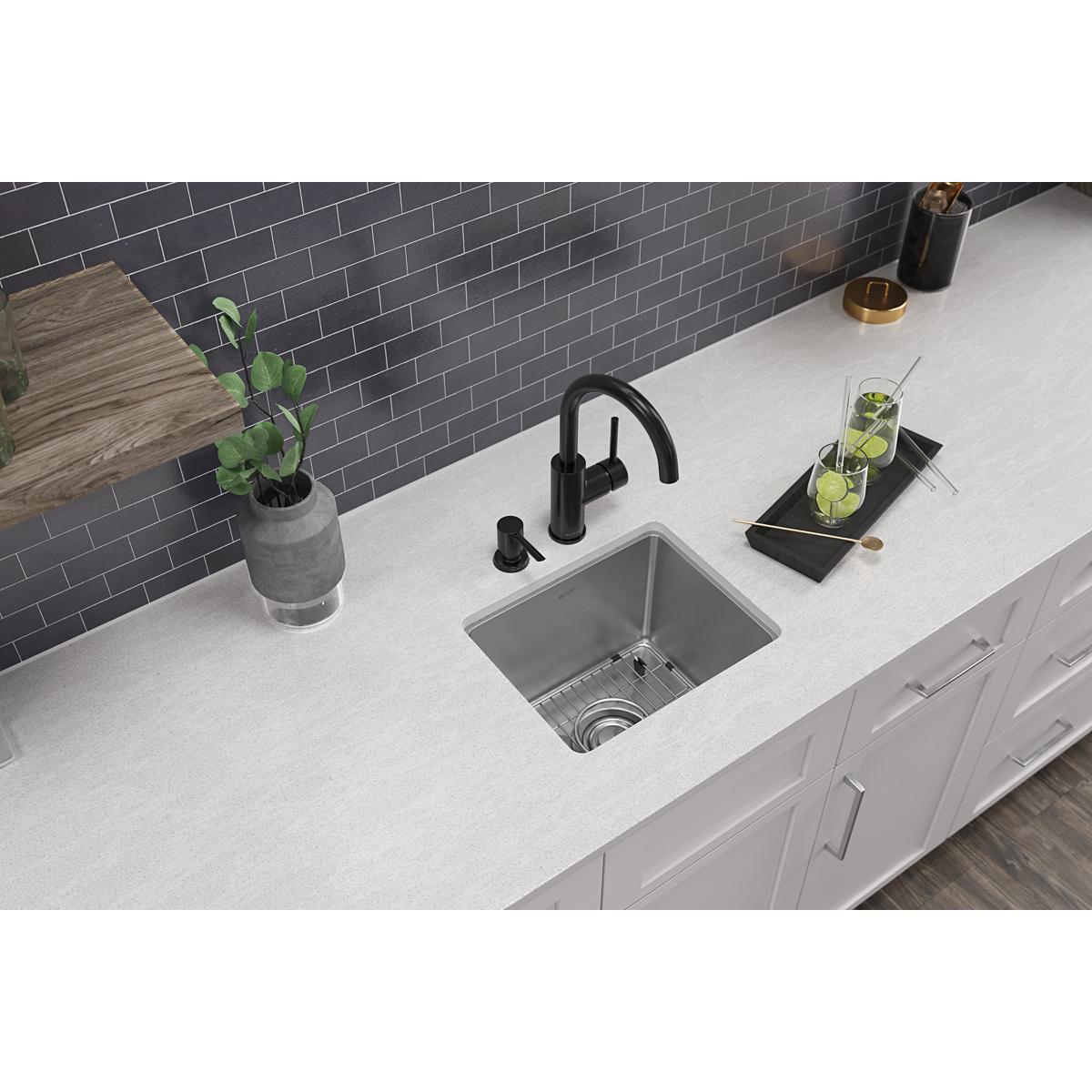 Stainless steel single bowl bar sink with two faucet holes mounted in white countertop with black faucet and accessories.