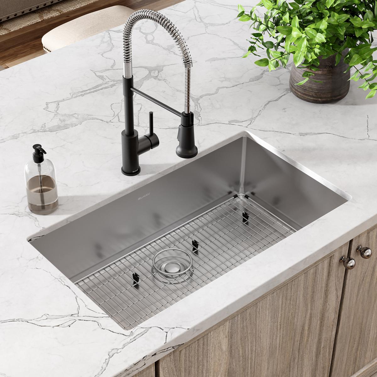 Stainless steel single bowl kitchen sink with grid and black pull-down faucet installed in marble countertop.