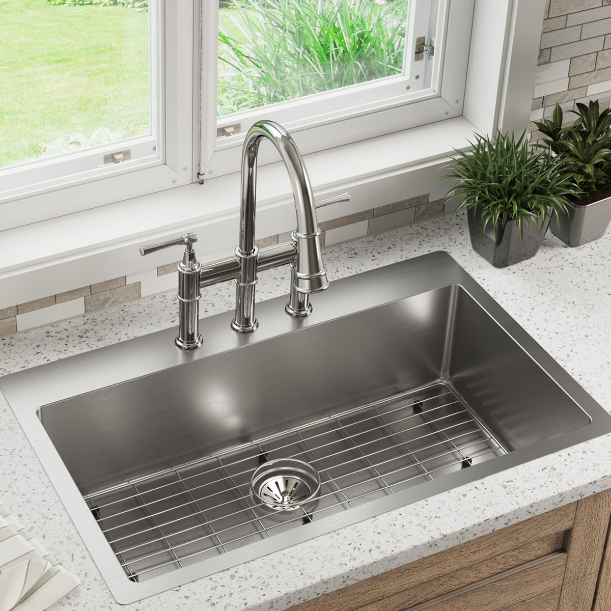 Stainless steel single bowl sink with four faucet holes, grid rack, and chrome bridge faucet installed in countertop.