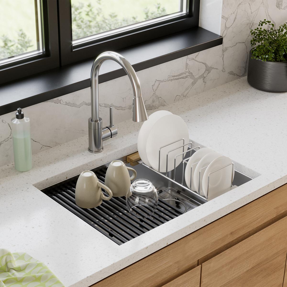 Stainless steel drying rack with wood handles positioned in kitchen sink next to white dishes and ceramic mugs.