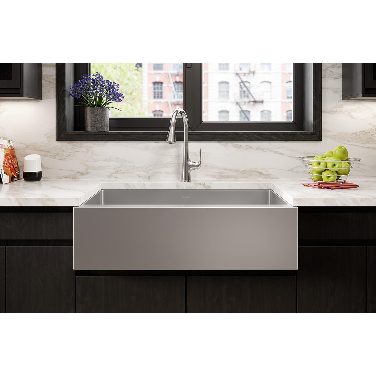 Stainless steel single bowl farmhouse sink with tall faucet installed in black cabinetry with marble countertop and backsplas