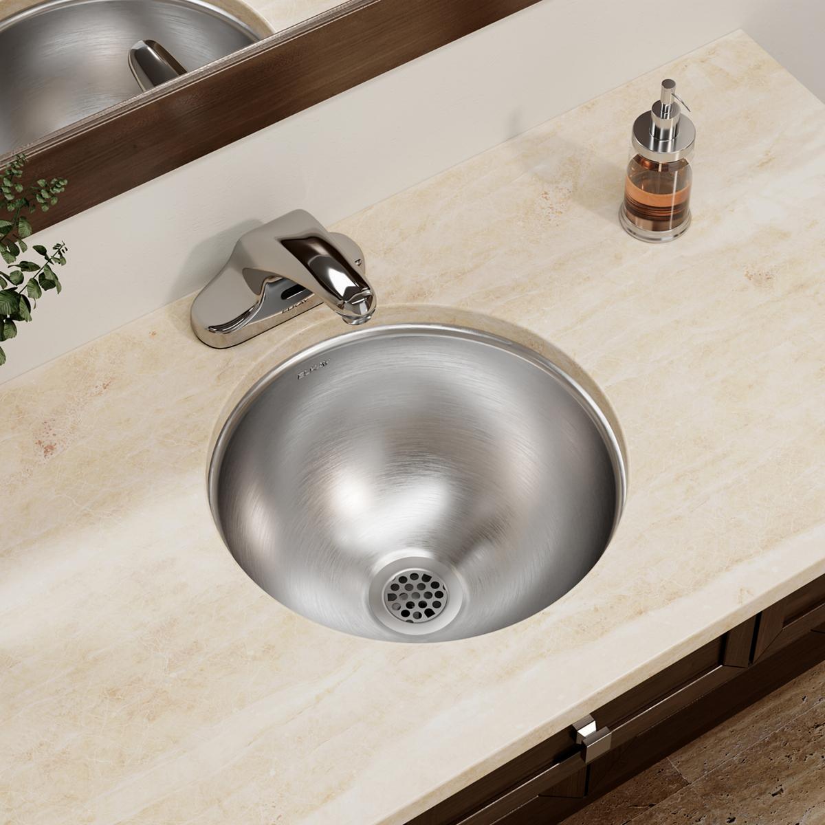Stainless steel round undermount bathroom sink installed in beige countertop with chrome faucet and soap dispenser.