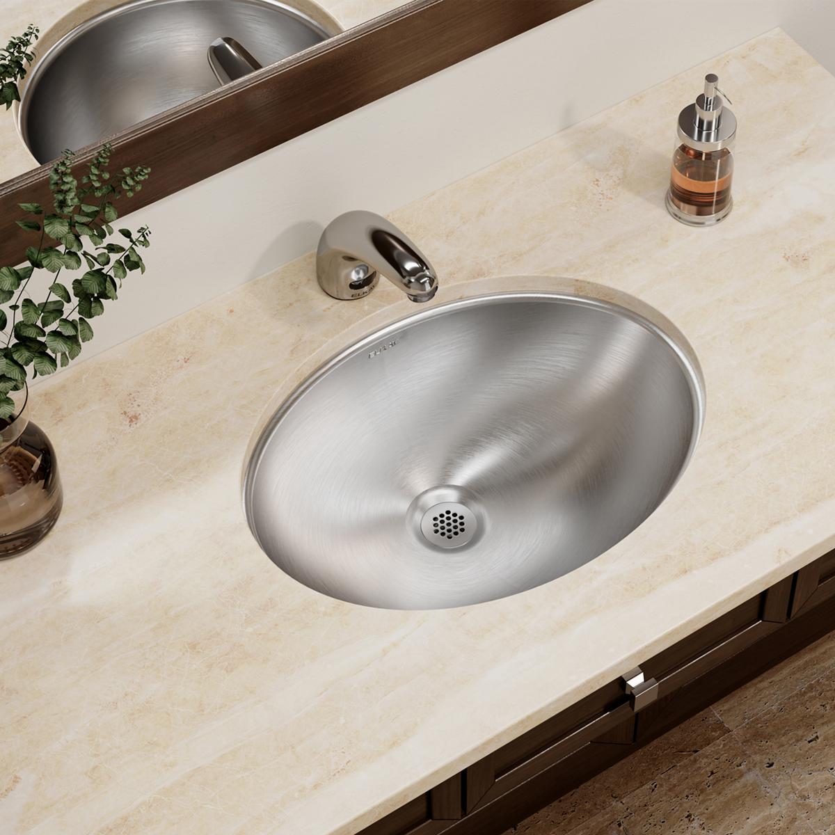 Stainless steel oval undermount bathroom sink installed in light stone countertop with faucet and accessories.