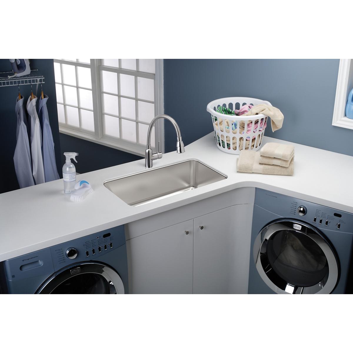 Stainless steel single bowl undermount sink installed in white countertop between two washing machines in laundry room.