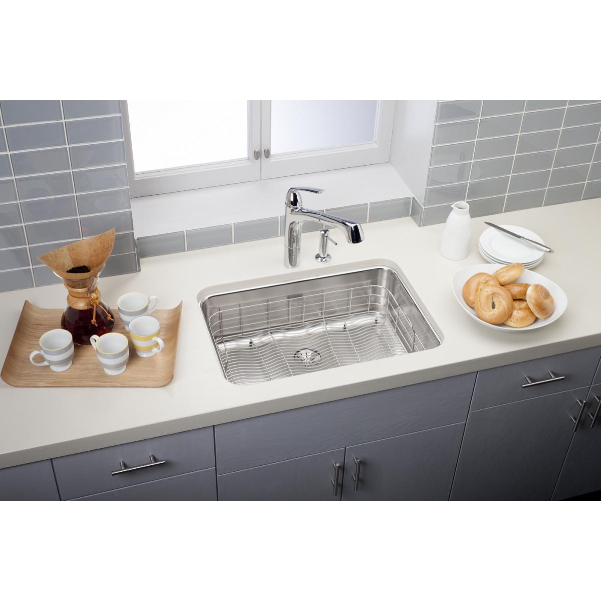 Stainless steel single bowl undermount sink with drain grid installed in white countertop next to chrome faucet.