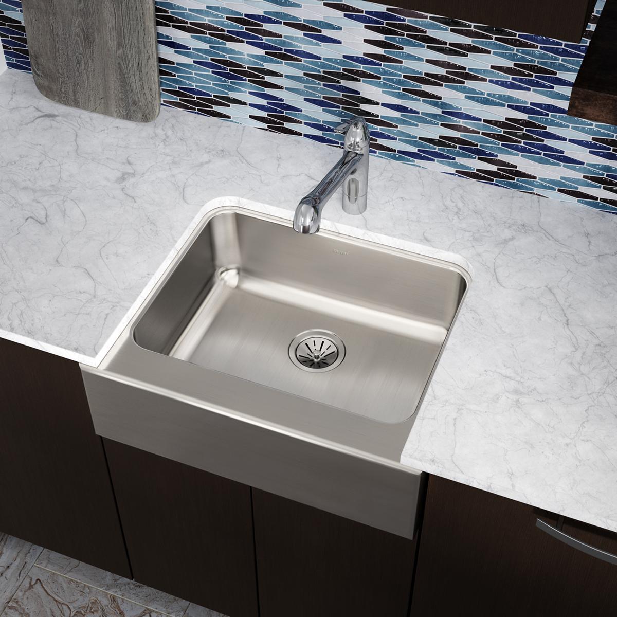 Stainless steel single bowl farmhouse sink installed in marble countertop with blue tile backsplash and chrome faucet.
