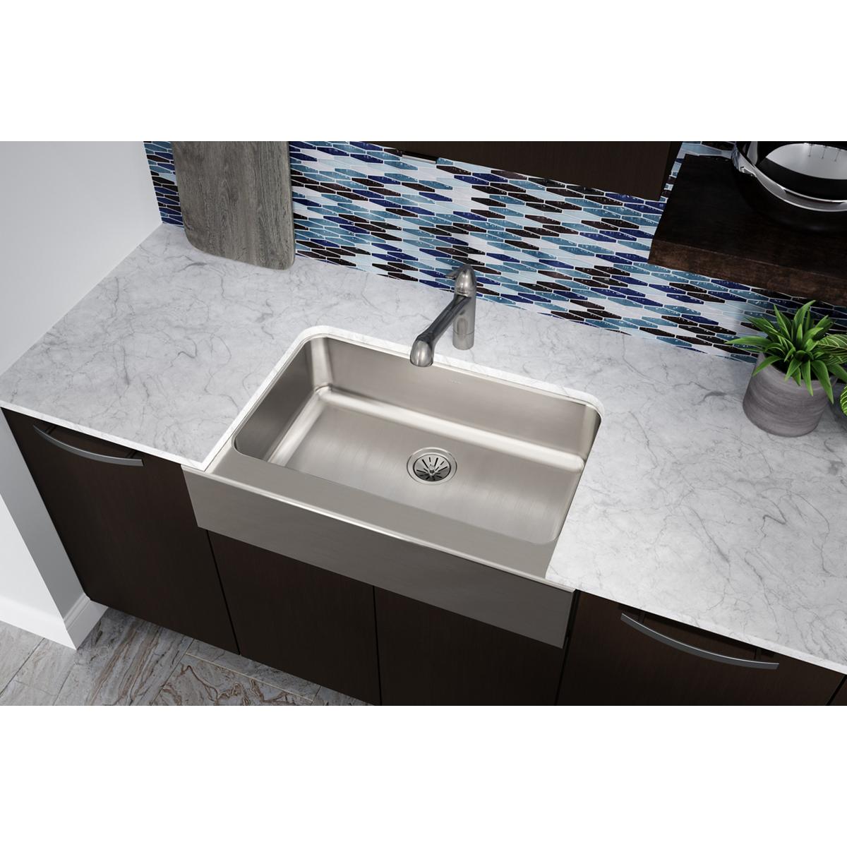 Stainless steel farmhouse sink with single large basin mounted on marble countertop with dark cabinetry below.