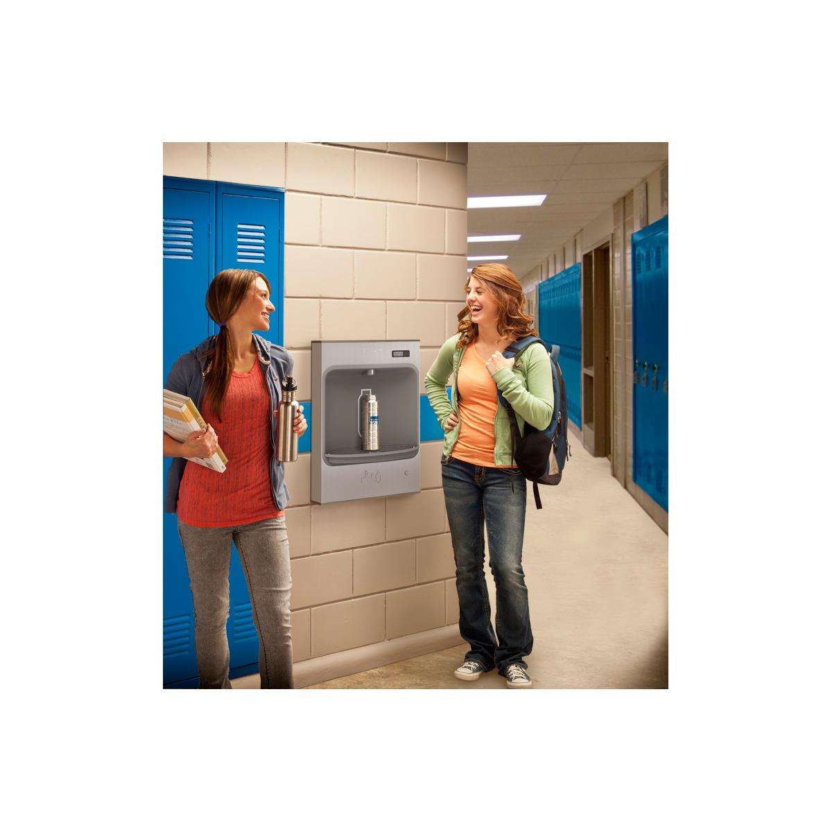 Stainless steel wall-mounted water bottle filling station installed in a school hallway with two students nearby.