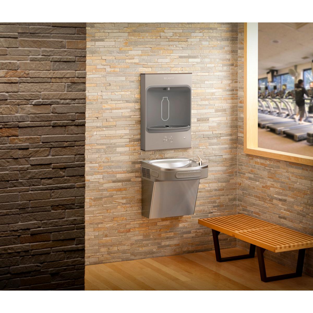 Stainless steel wall-mounted bottle filling station with mechanical controls mounted above a basin on brick wall.