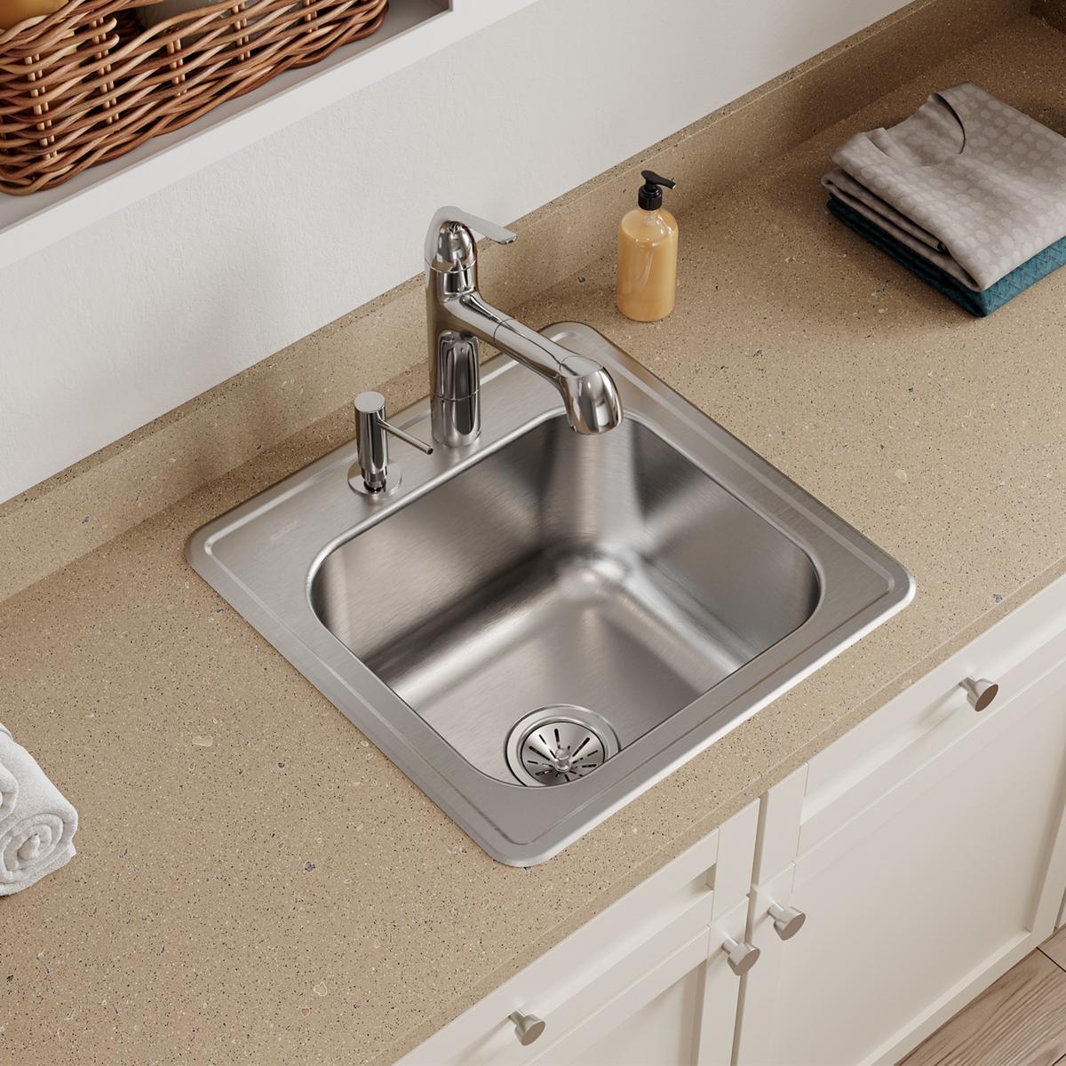 Stainless steel drop-in laundry sink with single bowl, two faucet holes, and drain installed in tan countertop.