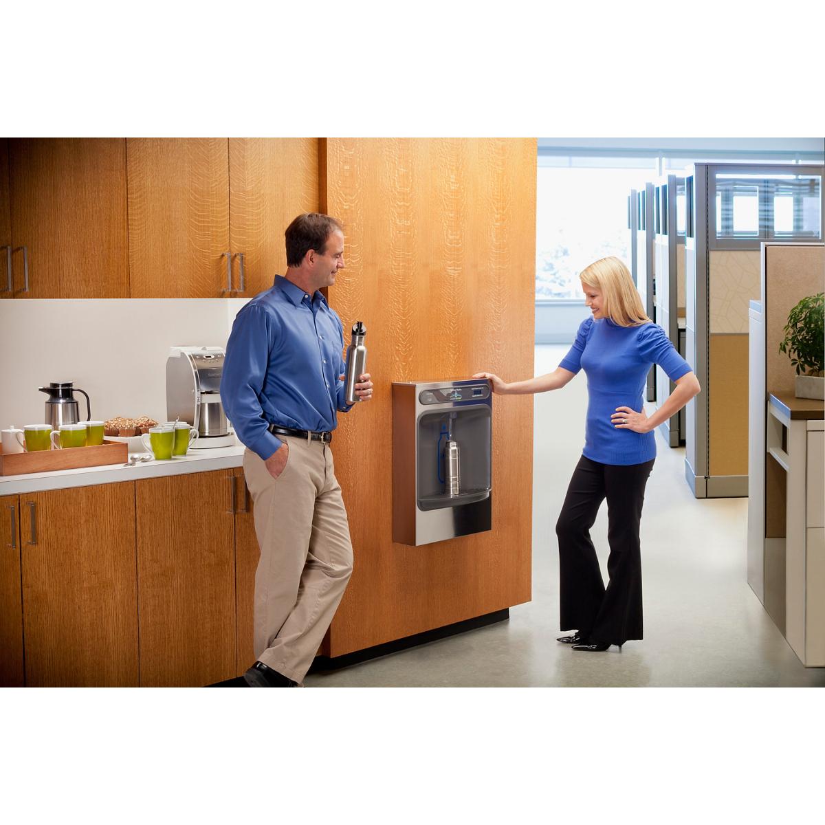 Stainless steel and dark gray water bottle filling station mounted on wooden wall panel in office kitchen area with two peopl