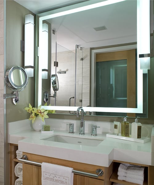 Rectangular lighted mirror with white frame mounted above bathroom sink with chrome faucet and white countertop.