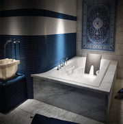 White rectangular drop-in soaking bathtub with raised deck, two armrests, and grab bars installed in marble surround.