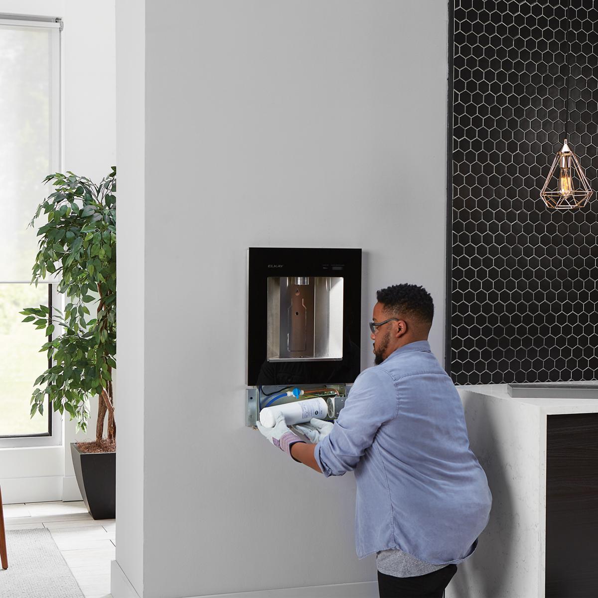 Man installing black wall-mounted water filter box with stainless steel interior and filter cartridge visible.