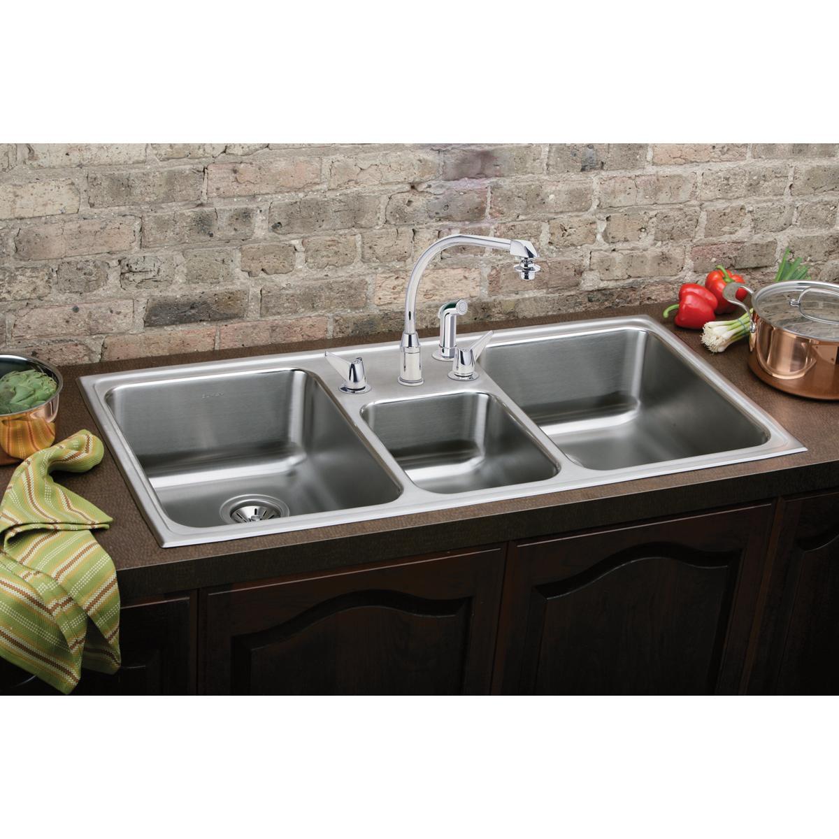 Stainless steel triple bowl drop-in sink with chrome faucet installed in dark cabinetry against brick backsplash.