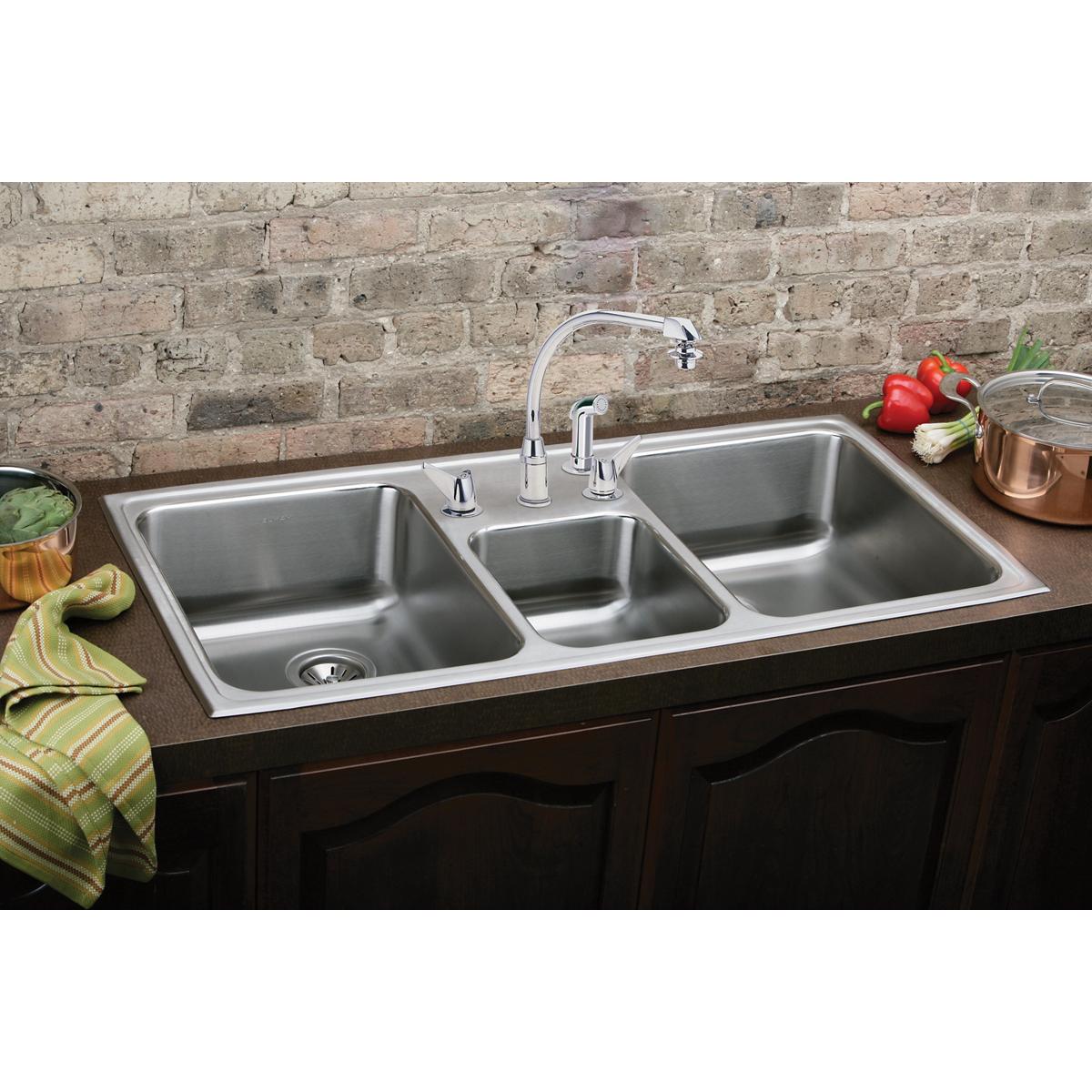 Stainless steel triple bowl drop-in sink with three faucet holes installed in countertop with brick backsplash.