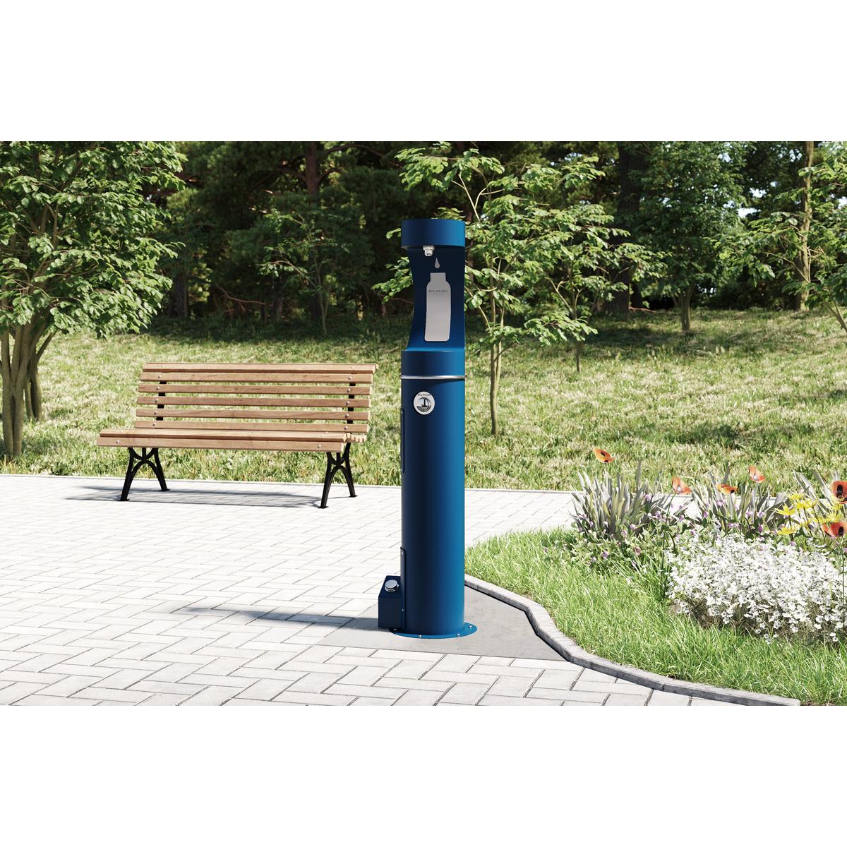 Blue outdoor bottle filler station with foot pedal control mounted on pavement near bench and garden.