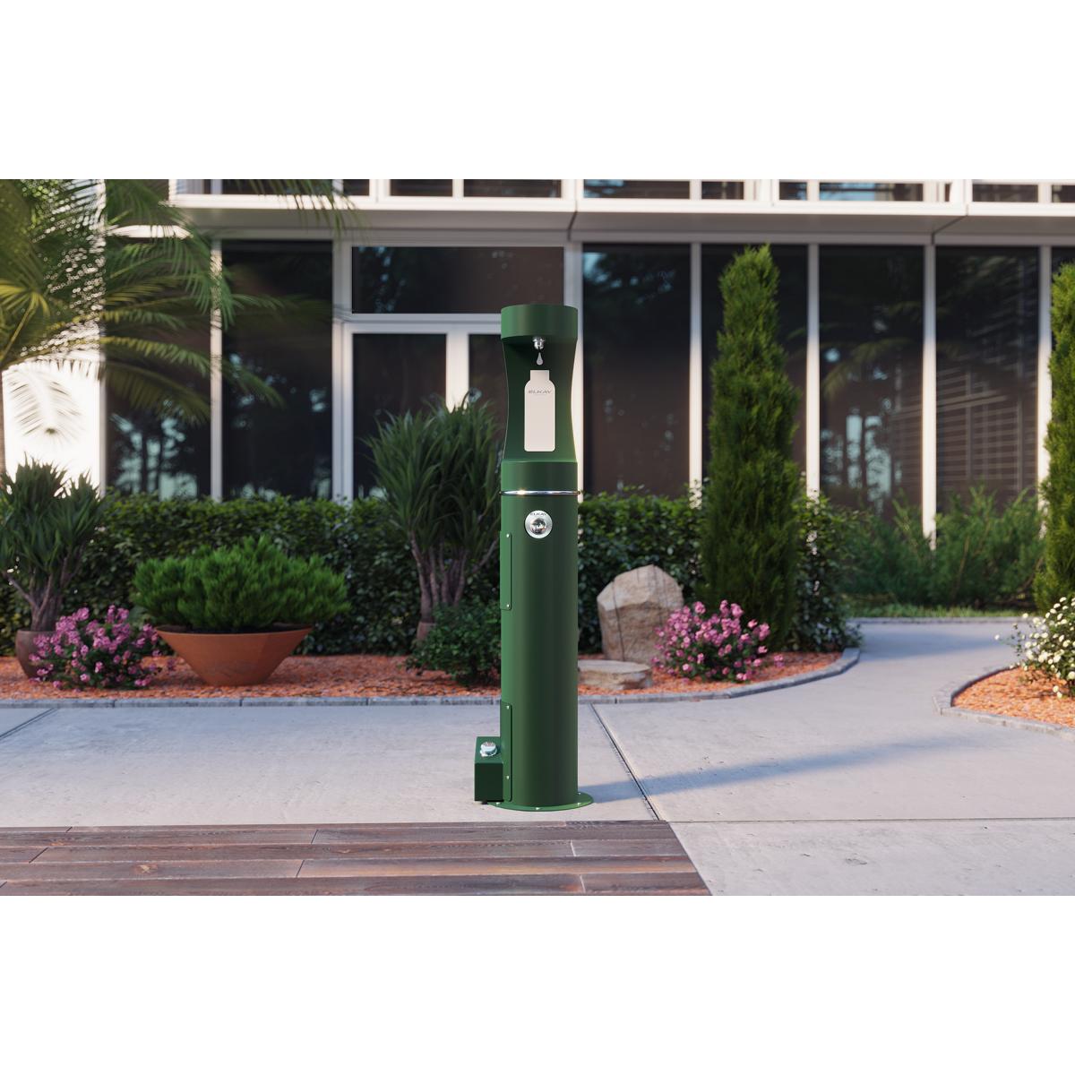Green outdoor bottle filler station with foot pedal base mounted on sidewalk in landscaped area.