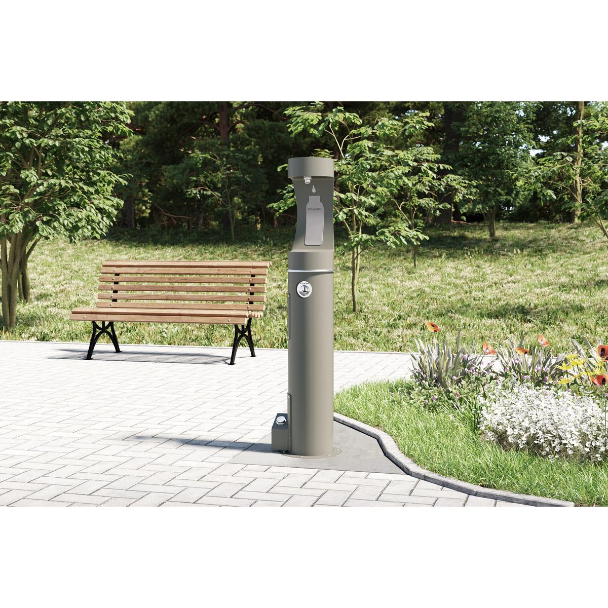 Gray outdoor bottle filler station with foot pedal control mounted on a paved surface near a wooden bench.