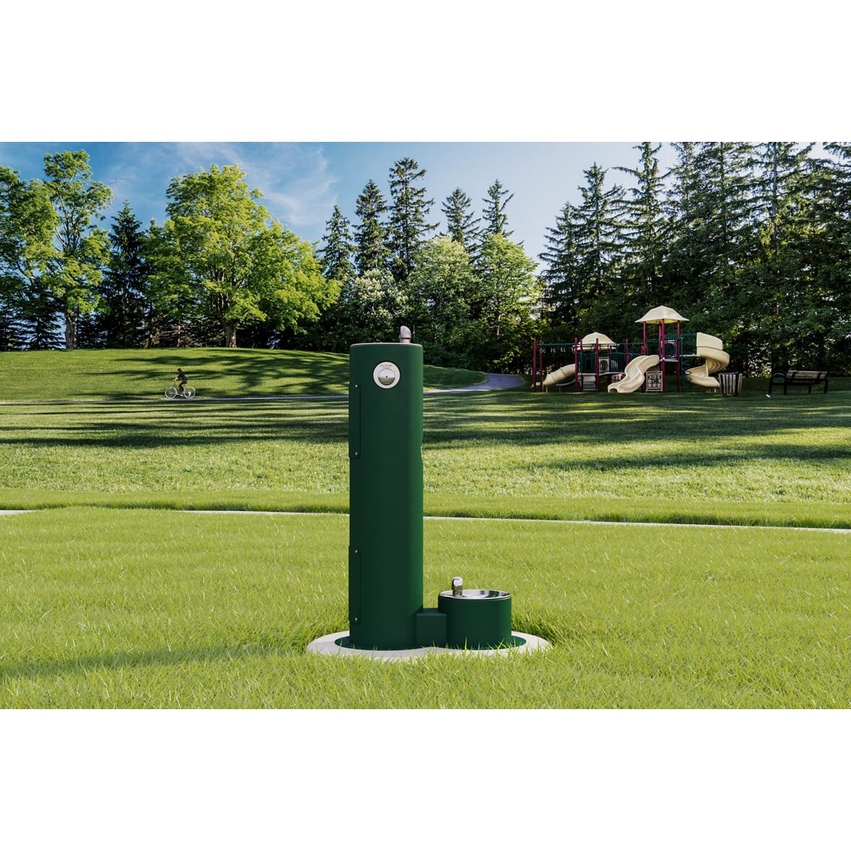 Green pedestal fountain with lower pet bowl station installed in a park setting with trees and playground in background.