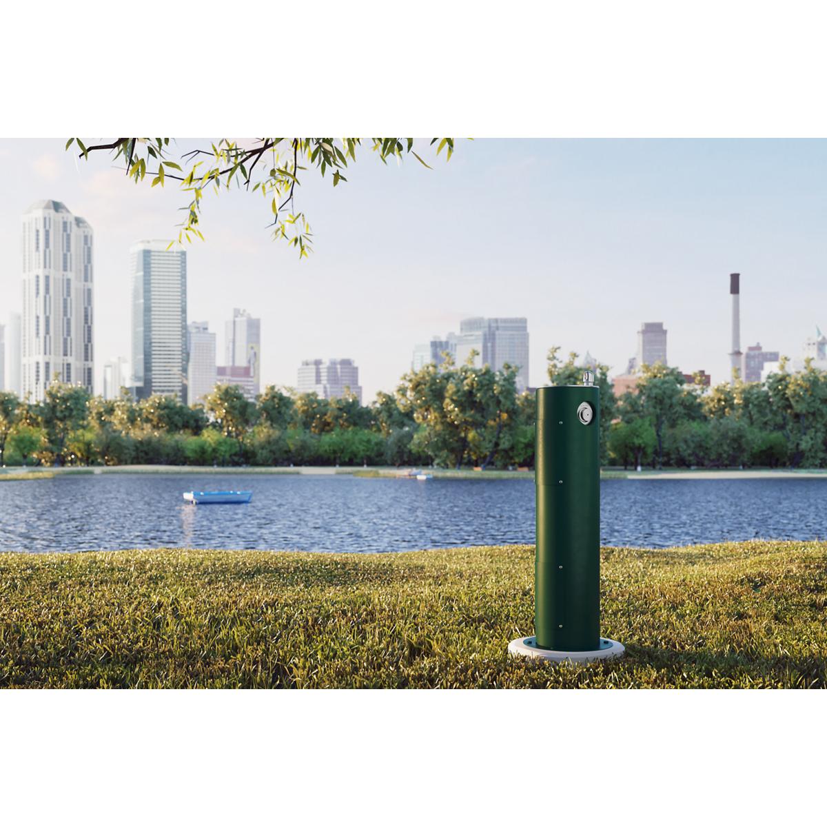 Tall green pedestal fountain with white base installed on grass near water with city skyline in background.