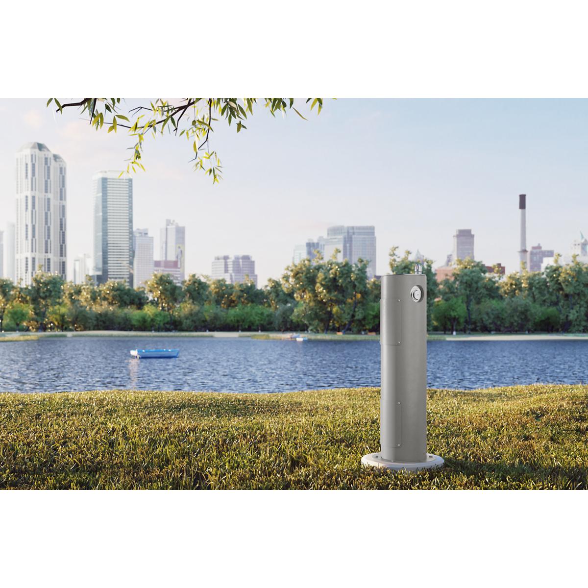 Gray cylindrical pedestal fountain standing on grass near water with city skyline in background.