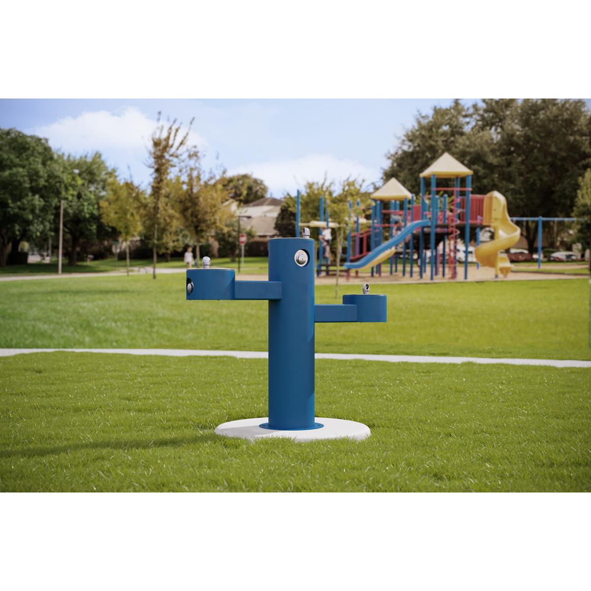 Blue pedestal drinking fountain with three staggered bowl levels mounted on concrete base in outdoor park setting.