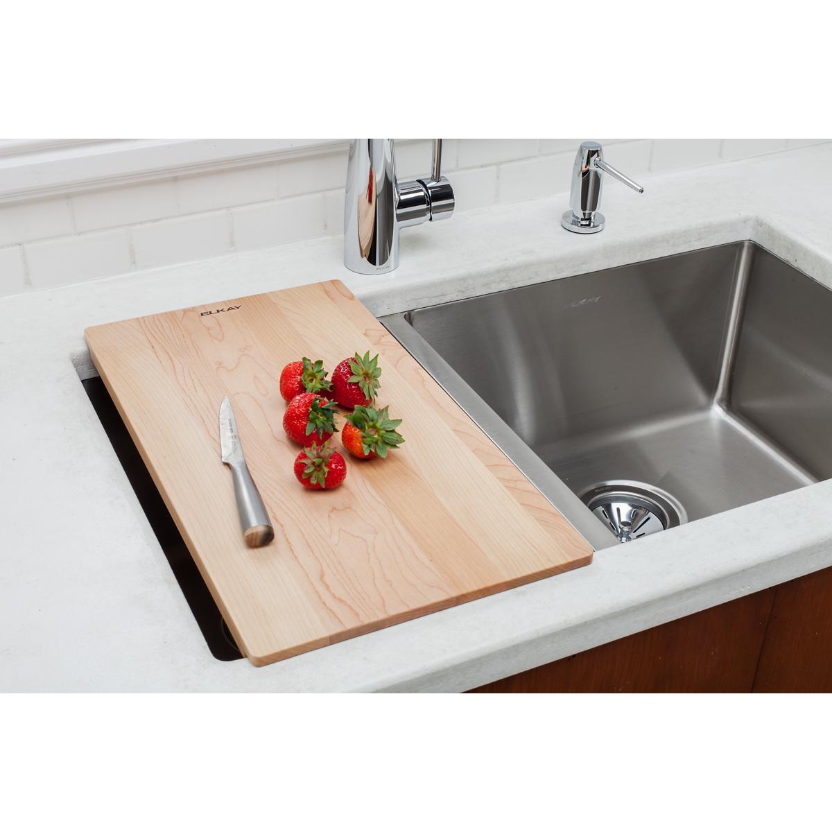 Light wood cutting board positioned over kitchen sink with fresh strawberries and knife on surface.