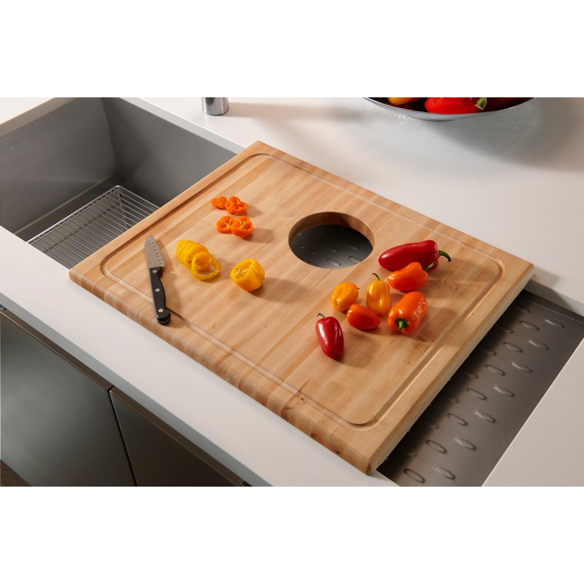 Hardwood cutting board with rounded edges and center hole, positioned over kitchen sink with chopped peppers and knife.