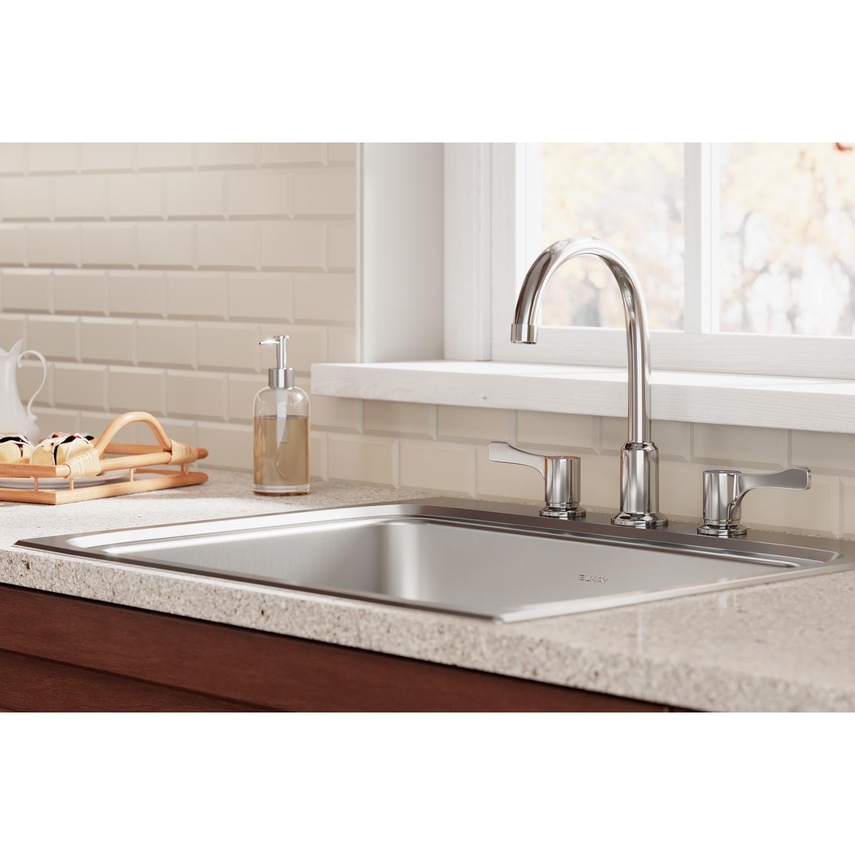 Chrome faucet with curved spout and lever handles installed on kitchen sink with white subway tile backsplash.