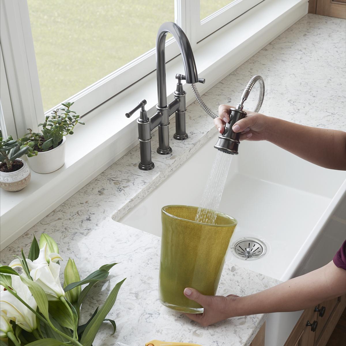 Antique steel bridge faucet with pull-down spray and lever handles installed over white sink with marble countertop.