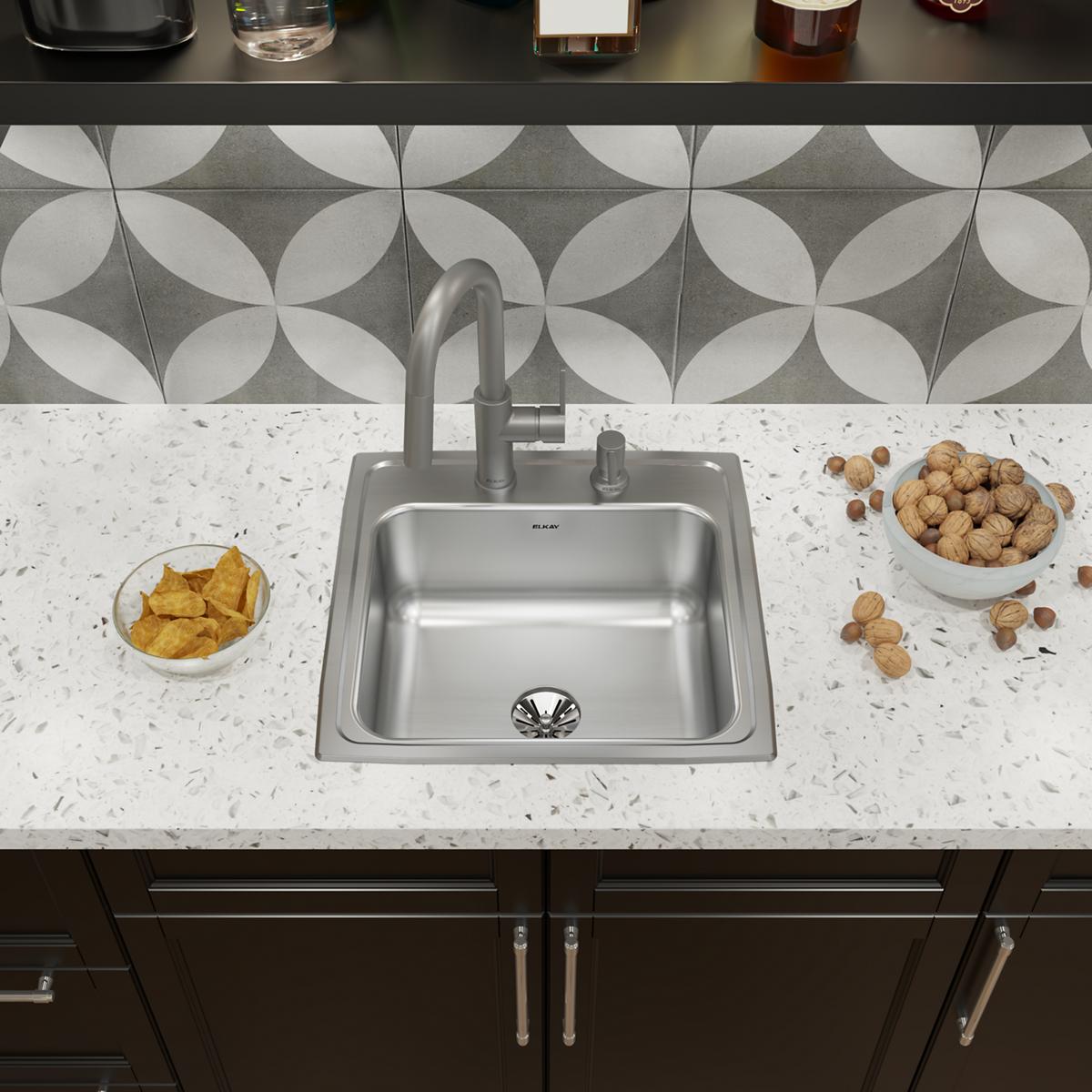 Stainless steel drop-in kitchen sink with single bowl, two faucet holes, and drain installed in white countertop.