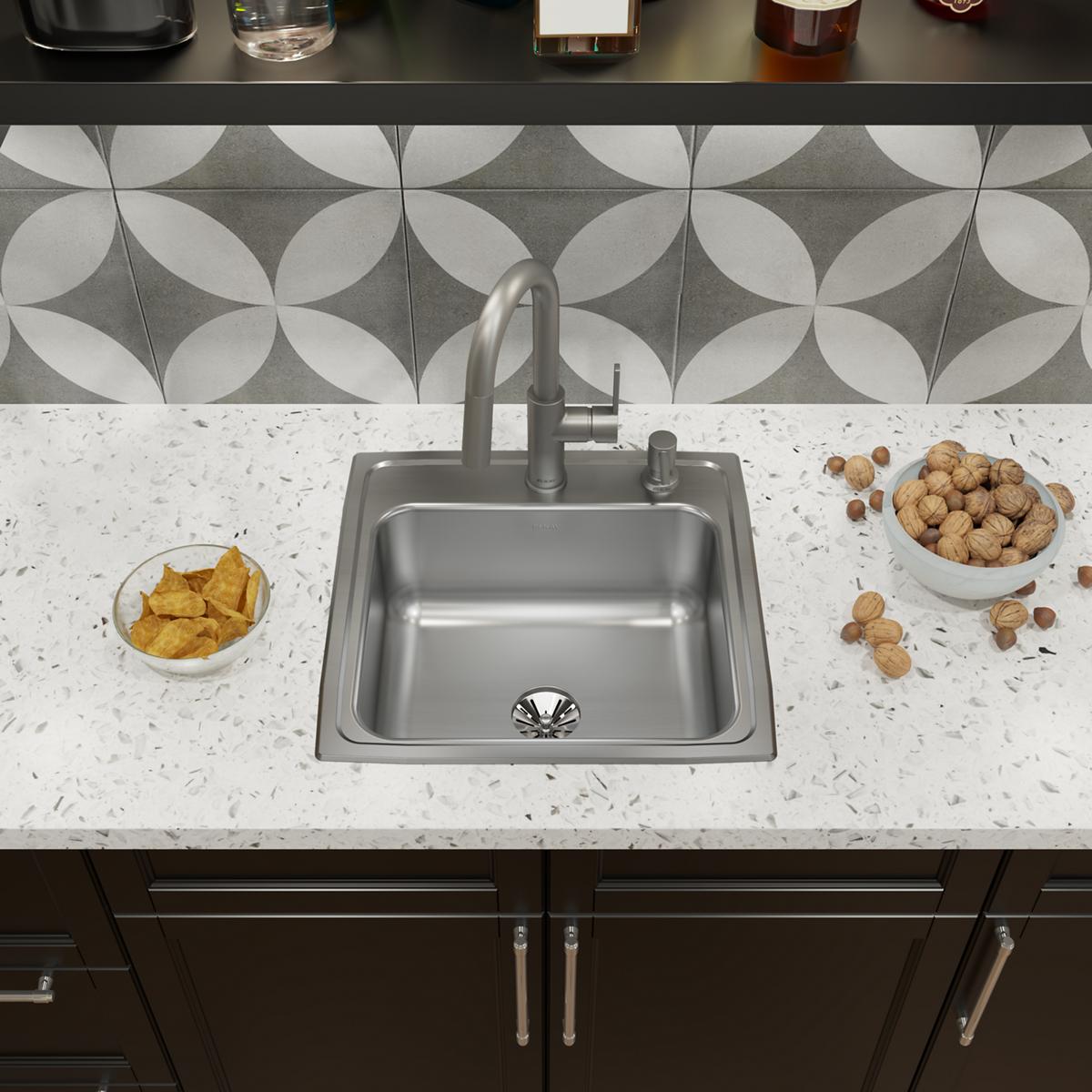 Stainless steel drop-in kitchen sink with single bowl, faucet, and drain installed in white speckled countertop.