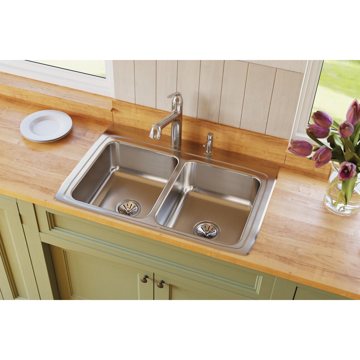 Stainless steel double bowl drop-in sink with two faucet holes installed in wood countertop above green cabinetry.