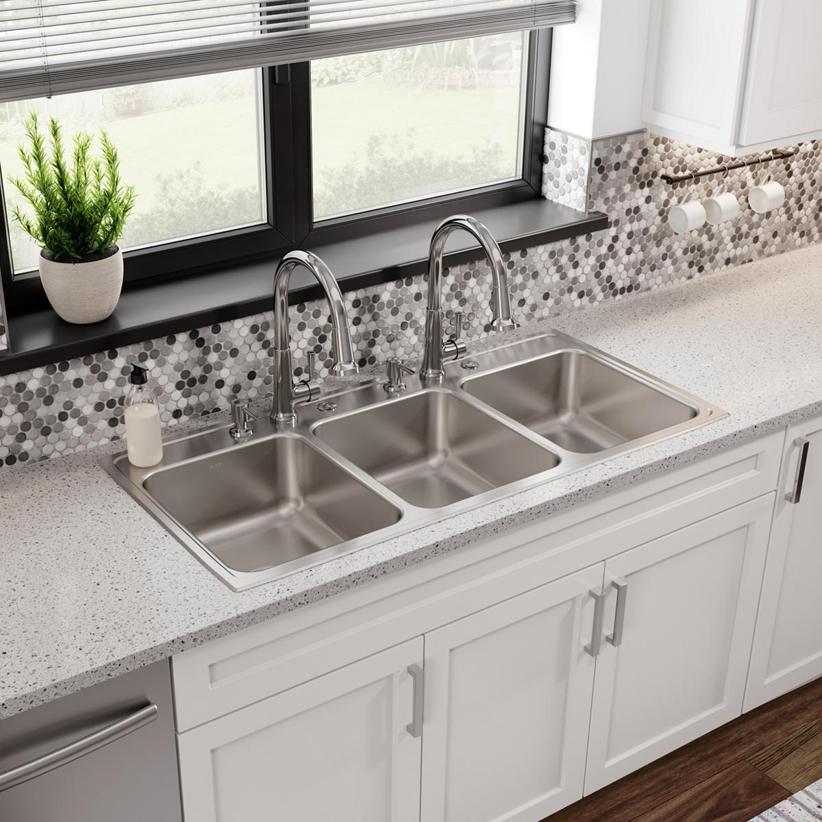 Stainless steel triple bowl drop-in sink with two chrome faucets installed in white kitchen countertop.