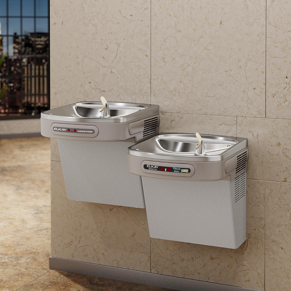 Light gray Elkay dual-level water cooler with stainless steel bowls and hands-free activation mounted on beige tile wall.