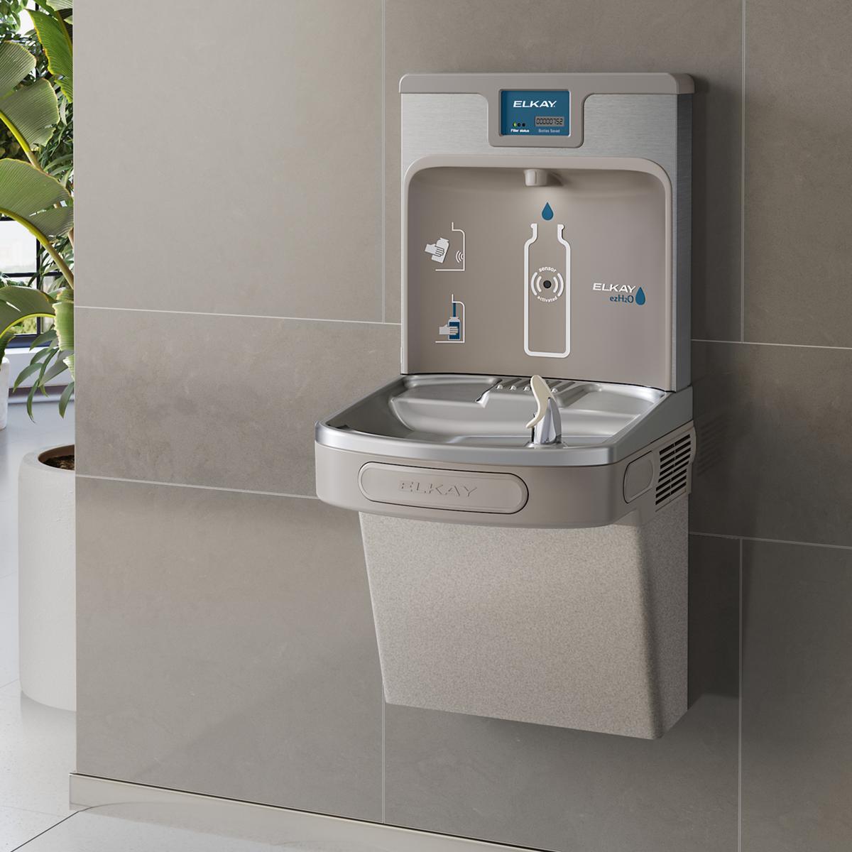 Light gray Elkay water fountain and bottle filling station with curved basin and control panel mounted on wall.