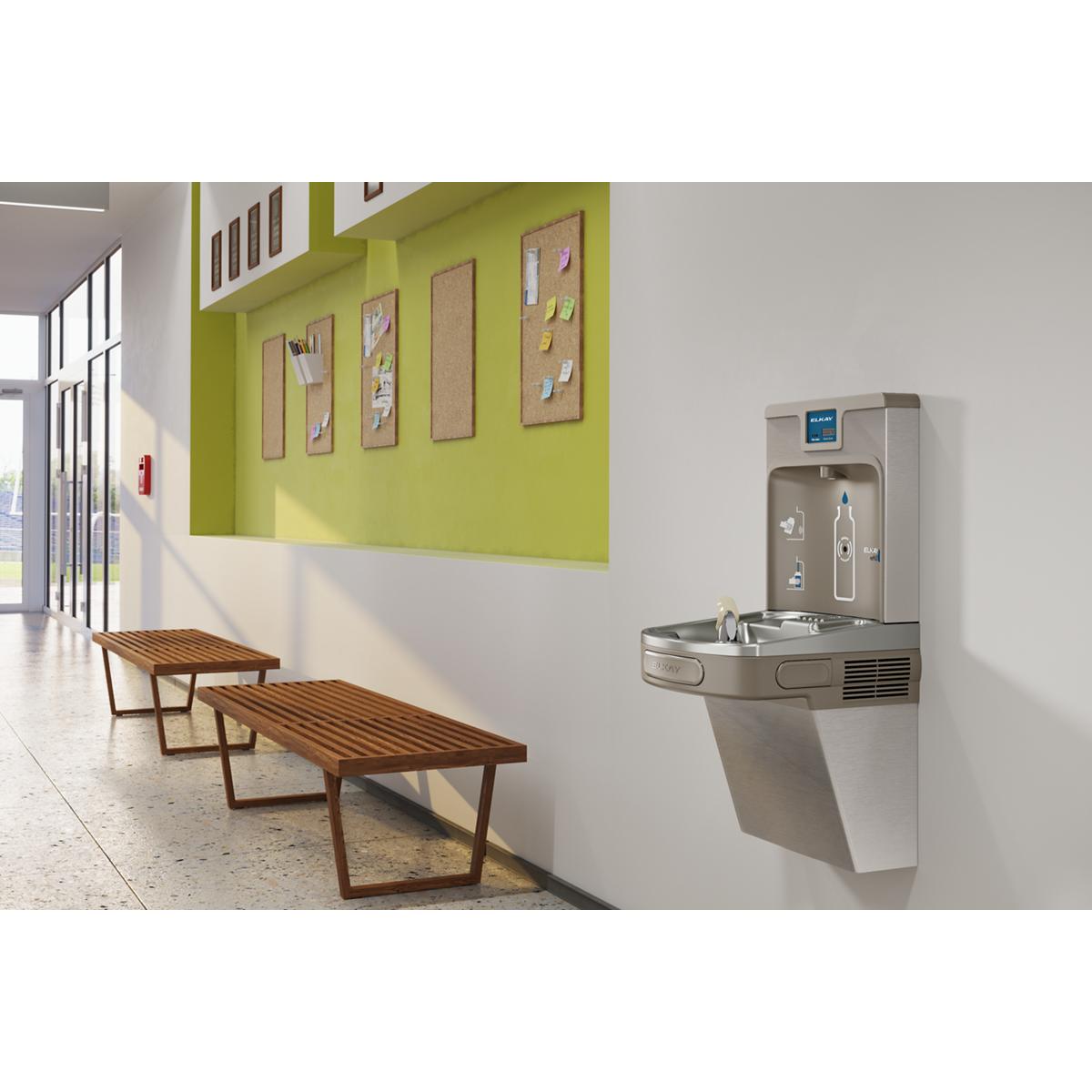 Stainless steel wall-mounted water cooler and bottle filling station in institutional hallway with benches.