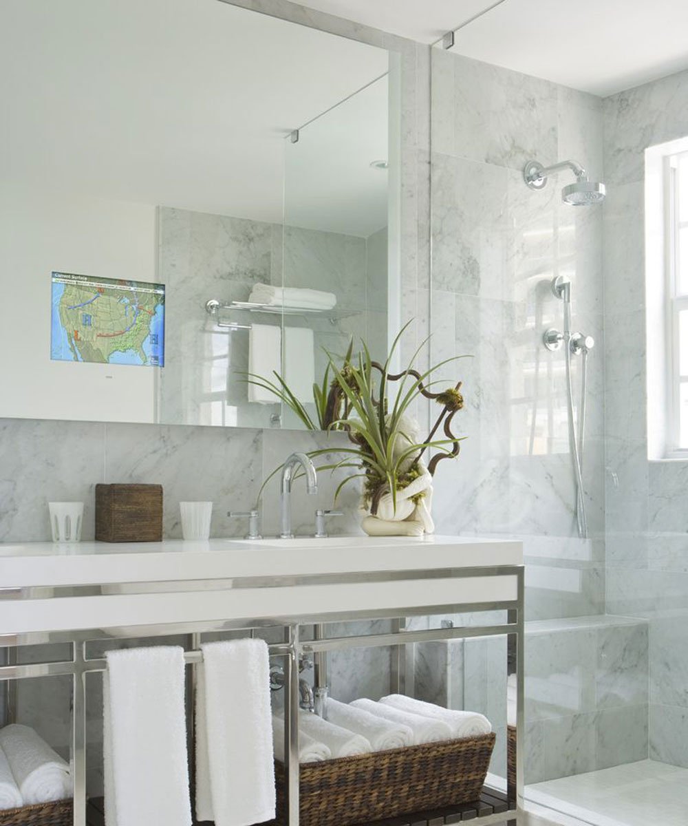 Marble bathroom with large mirror featuring embedded TV displaying a map, above white vanity with plant and accessories.
