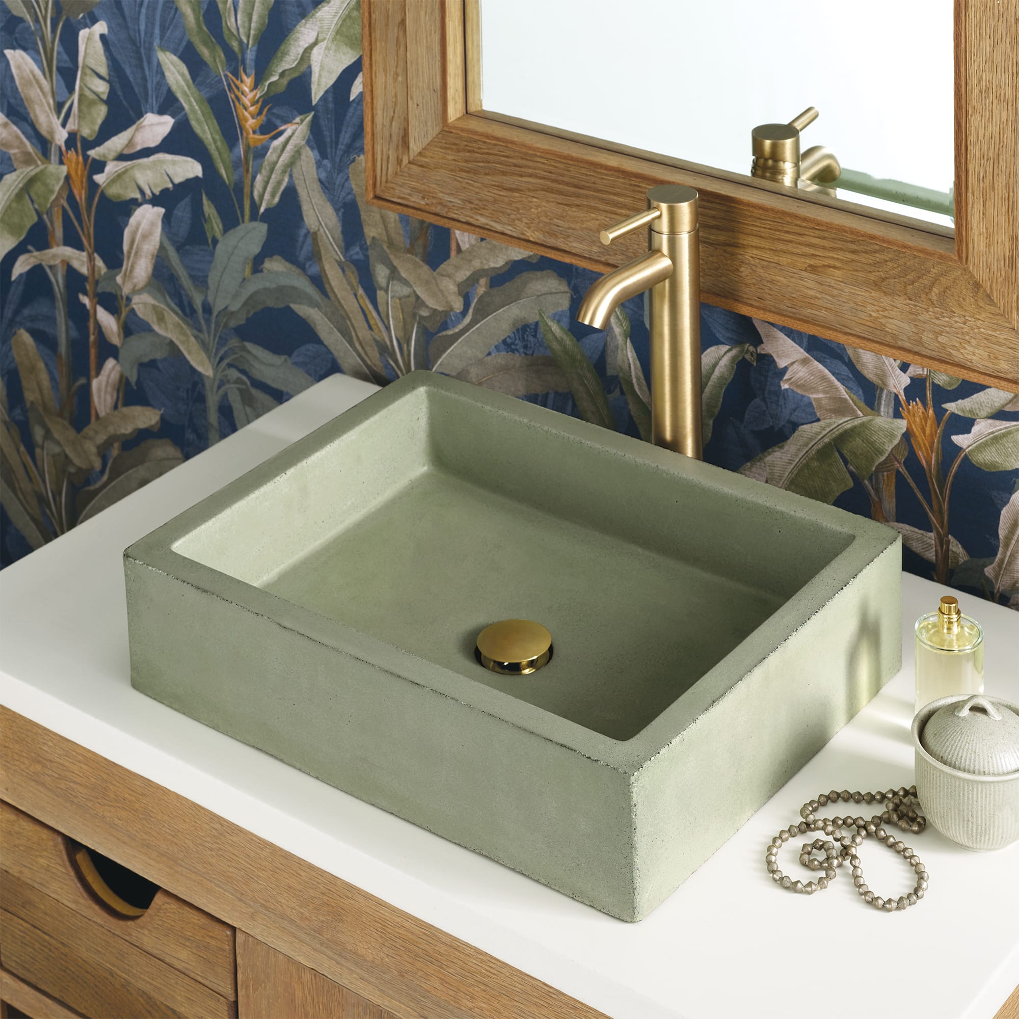 Sage green rectangular concrete sink with brass faucet on wooden vanity with botanical wallpaper backdrop.