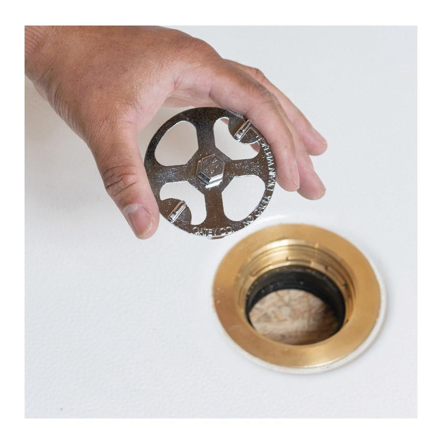Hand holding dark metal wrench with four circular openings above brass shower drain fitting.
