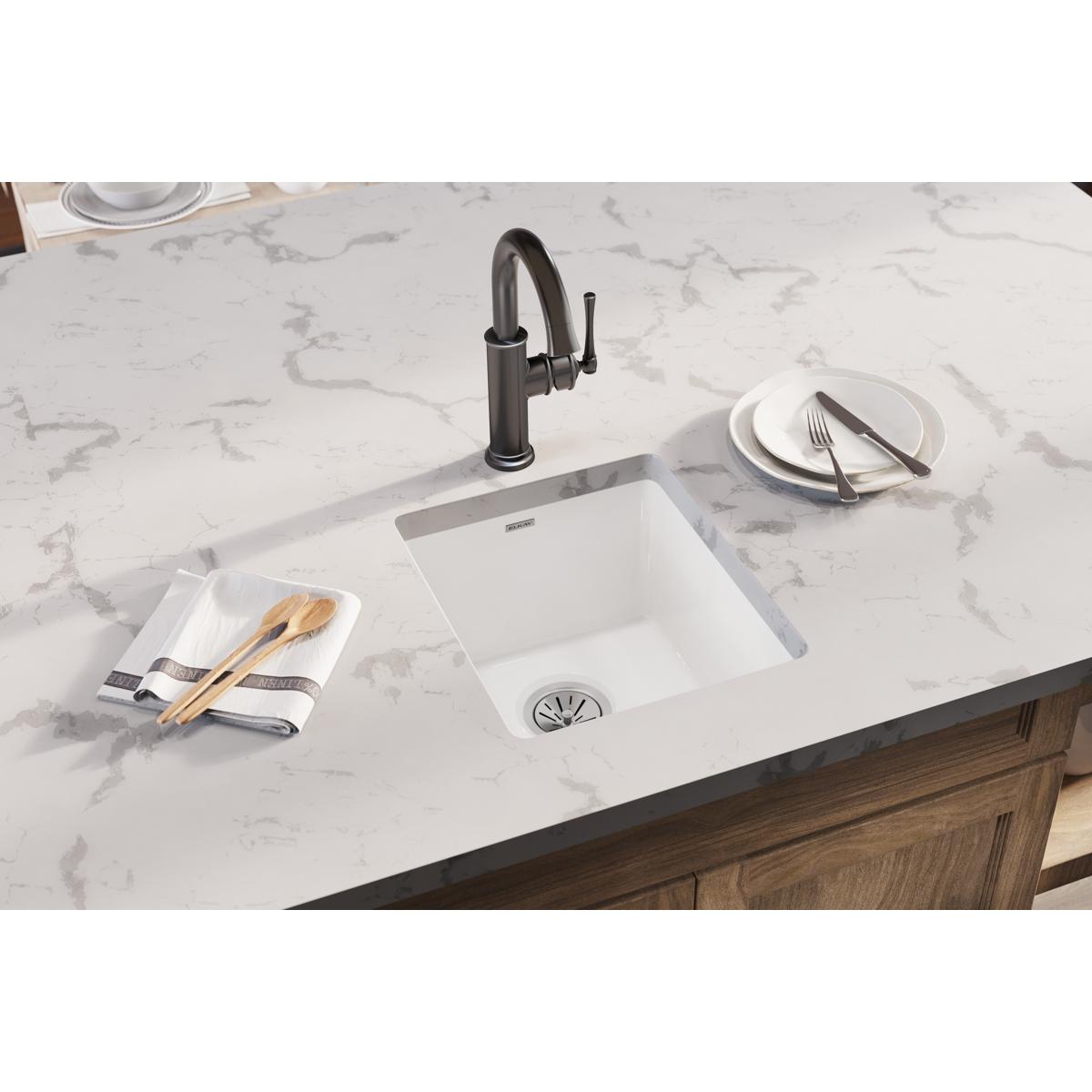 White fireclay undermount bar sink installed in marble countertop with black faucet.