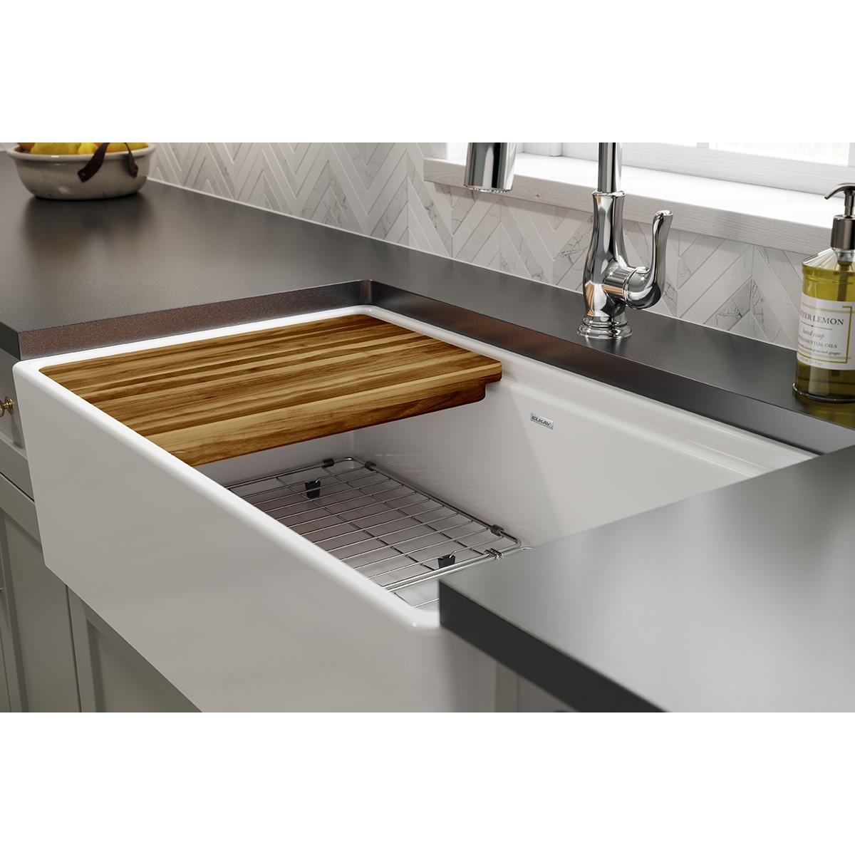 Hardwood cutting board with grain pattern resting over white farmhouse sink with chrome faucet.
