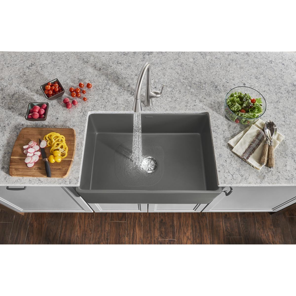 Gray fireclay single bowl farmhouse sink installed in countertop with faucet, surrounding fresh produce and kitchen items.